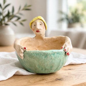 May include: A ceramic bowl featuring a sculpted figure with a yellow cap, red lips, and red fingernails. The bowl has a textured, light green exterior and a speckled cream interior, with the figure's arms wrapped around the sides of the vessel.