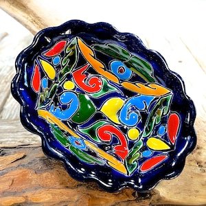 Könnte beinhalten: Eine blaue Keramikschale mit gewelltem Rand und einem farbenfrohen, handbemalten Design. Das Design zeigt Wirbel, Blätter und geometrische Formen in Rot-, Gelb-, Grün- und Blautönen.
