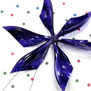 Pu&ograve; includere: Un fiocco metallico viola a sei punte, che ricorda una stella, su uno sfondo bianco con pois colorati.