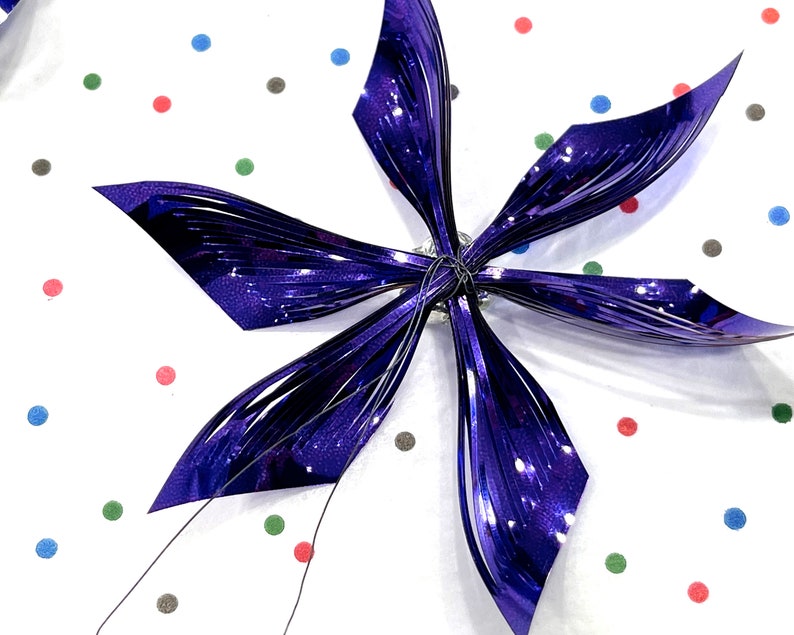 Pu&ograve; includere: Un fiocco metallico viola a sei punte, che ricorda una stella, su uno sfondo bianco con pois colorati.