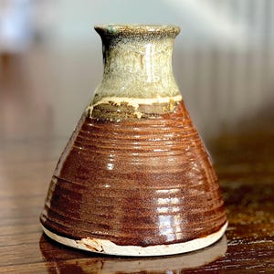 May include: A ceramic vase with a wide, conical base and a narrow neck. The lower portion features a glossy, dark brown glaze with horizontal ridged textures, while the upper neck is finished in a contrasting, mottled light beige and grey glaze.