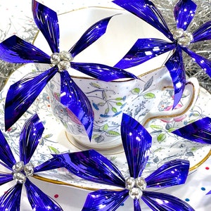Pu&ograve; includere: Una tazza da t&egrave; e un piattino bianchi con un motivo floreale, decorati con quattro fiocchi blu iridescenti con centri gioiello.