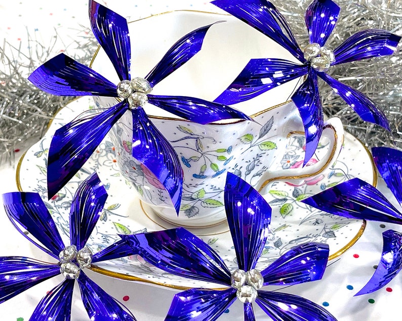 Pu&ograve; includere: Una tazza da t&egrave; e un piattino bianchi con un motivo floreale, decorati con quattro fiocchi blu iridescenti con centri gioiello.