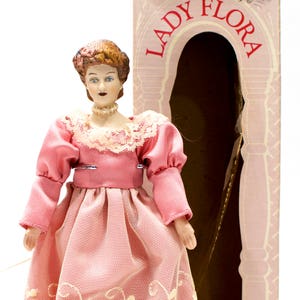 Puede incluir: Una muñeca de porcelana con un vestido rosa con ribetes de encaje y un collar de perlas. La muñeca está en una caja con las palabras "Lady Flora" impresas en ella.