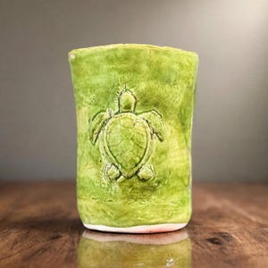 May include: A cylindrical ceramic cup with a vibrant lime green glaze and a textured surface. The exterior features a prominent, raised relief sculpture of a sea turtle centered on the front, showcasing detailed shell patterns and flippers.