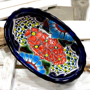 Op de afbeelding: Een ovale keramische serveerschaal met een handgeschilderd bloemenmotief. De schaal is blauw en wit met rode, gele, blauwe en groene bloemen op een witte achtergrond. De rand van de schaal is donkerblauw.