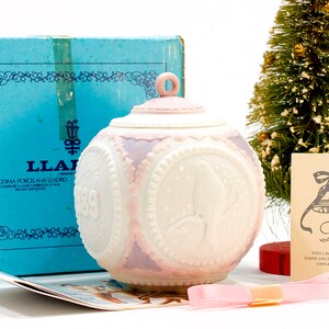 May include: A white porcelain Christmas bauble with a pink and purple design. The bauble features a dove with a snowflake pattern and the year 1989. The bauble is in its original box with a blue and white label that reads "Legitima Porcelana Lladro".