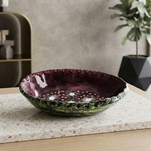 May include: A shallow, round ceramic bowl with a glossy, deep plum-colored interior featuring a series of white, star-shaped floral patterns. The exterior of the bowl has a textured, dark green finish with a scalloped, wavy rim edge.