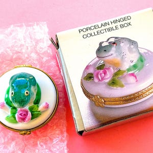 Pode incluir: Caixa colecionável de porcelana com dobradiças e uma figura de sapo. O sapo é verde e azul, com rosas cor-de-rosa e folhas verdes. A caixa é branca com detalhes dourados. Um item colecionável.