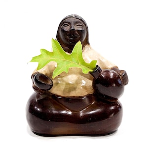 Puede incluir: Una figurita de cerámica de una persona sentada con las piernas cruzadas, sosteniendo una hoja y una olla pequeña. La figurita es marrón y blanca con una cara sonriente.