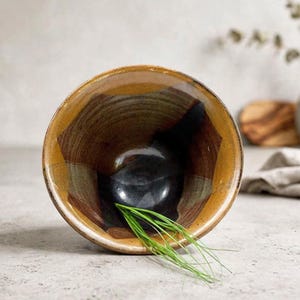 May include: A round ceramic bowl with a wide opening, featuring a glossy interior glaze in shades of deep black, warm brown, and golden ochre. The bowl has a smooth, curved shape and contains a small cluster of fresh green chives at the bottom.