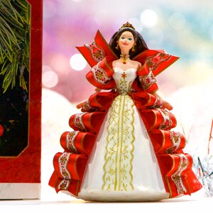 May include: A Christmas ornament in the shape of a doll, featuring a red and white gown with gold accents. The doll has brown hair, a crown, and is posed with arms outstretched. The ornament is part of a series, as indicated by the box in the background.