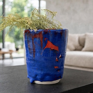 May include: A cylindrical ceramic planter with a vibrant blue glaze and visible drips of deep red and orange running down the sides. The surface has an uneven, textured finish with a white base rim, containing a sprig of dried green foliage.