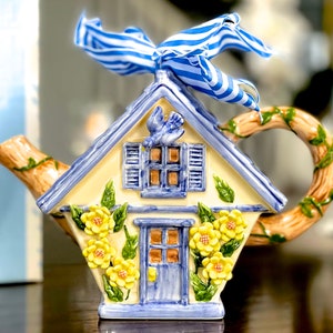 May include: A blue and yellow ceramic teapot shaped like a cottage with a blue bird on the front and a blue and white striped ribbon on the top. The teapot has a brown vine-like handle.