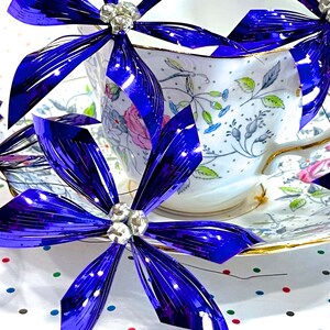 Pu&ograve; includere: Una tazza da t&egrave; e un piattino bianchi con un motivo floreale, decorati con fiocchi metallici blu e argentati. I fiocchi sono disposti intorno alla tazza da t&egrave; e al piattino, creando un look festoso ed elegante.