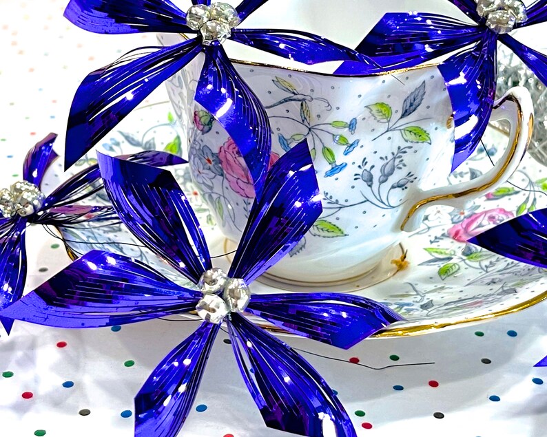 Pu&ograve; includere: Una tazza da t&egrave; e un piattino bianchi con un motivo floreale, decorati con fiocchi metallici blu e argentati. I fiocchi sono disposti intorno alla tazza da t&egrave; e al piattino, creando un look festoso ed elegante.