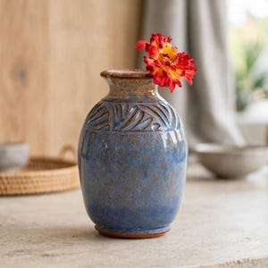 May include: A ceramic vase with a blue speckled glaze and a carved leaf pattern around the neck. The vase has a brown rim and base, and it contains a single bright red and yellow flower with layered petals extending from the opening.