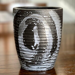May include: A cylindrical ceramic vessel with a dark charcoal gray base and a textured, ribbed surface. The exterior features a prominent, hand-painted white spiral design with speckled paint splatters that contrast against the dark background.