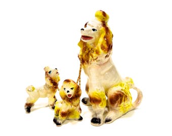May include: A set of three ceramic poodle figurines. The largest poodle is white with brown and yellow accents. The two smaller poodles are white with yellow accents. The poodles are all standing on a white background.
