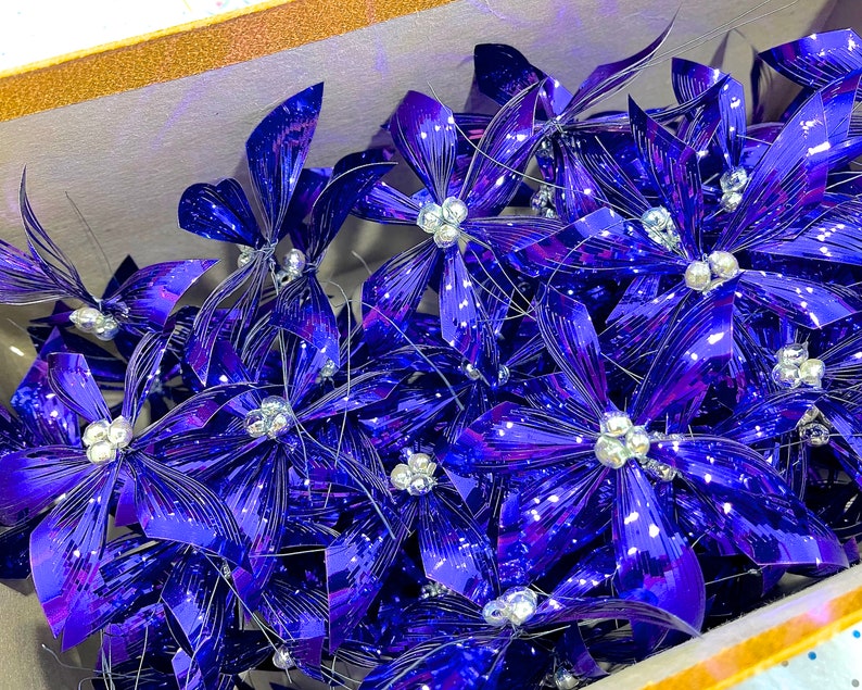 Pu&ograve; includere: Un mazzo di fiocchi metallici blu con centri argentati. I fiocchi sono disposti in una scatola e sono pronti per essere utilizzati per decorare.