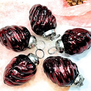 May include: Five dark red, ribbed, oval-shaped glass ornaments with a metallic finish. Each ornament features a silver-colored metal cap at the top with a small circular loop for hanging, arranged in a circular pattern on a light surface.