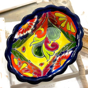 Pode incluir: Um pequeno prato de cerâmica oval com uma borda ondulada. O prato é pintado com um fundo amarelo brilhante e desenhos florais coloridos em tons de vermelho, laranja, verde e azul.