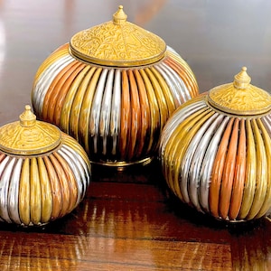 May include: Three decorative metal containers with lids. The containers are round and have a striped pattern of gold, silver, and copper. The lids are ornate and have a gold finish.