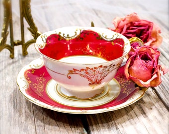 May include: A vintage teacup and saucer set with a red and gold floral design. The cup is filled with white liquid and the saucer has a scalloped edge.