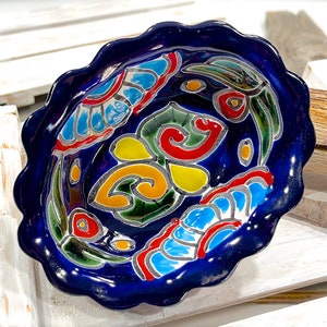 Könnte beinhalten: Eine blaue Keramikschüssel mit einem gewellten Rand und einem farbenfrohen Blumendesign. Die Schüssel zeigt gelbe, rote, grüne und blaue Blumen und Blätter.