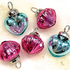 May include: Five small, vintage-style ornaments in the shape of hearts and rounded shapes. Two are teal, and three are magenta. Each has a silver-colored metal cap with a loop for hanging. The ornaments have a reflective, glass-like appearance.