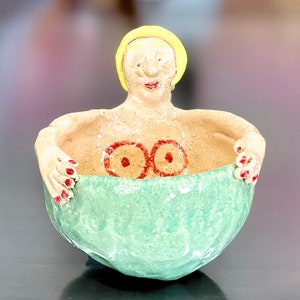 May include: A ceramic bowl shaped like a woman with a yellow head, red fingernails, and red painted breasts. The bowl is green on the inside and beige on the outside.
