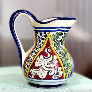 Pode incluir: Um jarro de cerâmica colorido com uma alça azul e um corpo branco decorado com padrões florais vermelhos, amarelos, verdes e azuis.