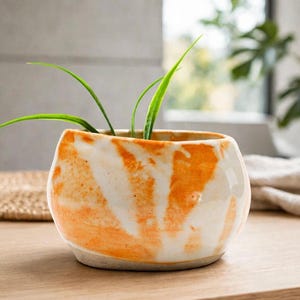May include: A small, round ceramic planter featuring a glossy white glaze with vibrant orange marbled streaks. The pot has a slightly irregular, organic shape and contains a green plant with thin, pointed leaves extending from the top opening.