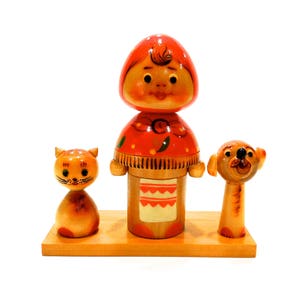 Pode incluir: Um conjunto de três figuras de madeira, uma menina com um vestido vermelho e branco, um gato e um cão. As figuras são pintadas com detalhes e têm um estilo de arte popular tradicional.