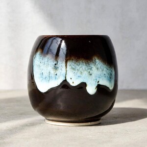 May include: A rounded ceramic cup featuring a glossy dark brown base glaze with a contrasting drip pattern of light blue and white glaze cascading from the rim. The vessel has a smooth, curved silhouette and a circular base with a light rim.