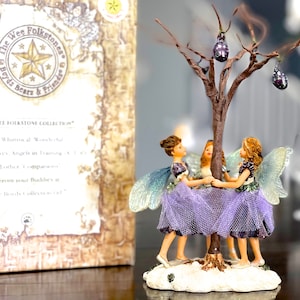 May include: Three fairies in purple dresses and wings are holding hands around a small tree with purple ornaments. The fairies are standing on a snowy base. The background is a book with the text "The Wee Folkstones Boyds Bears & Friends".