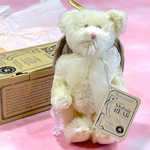 May include: A white mohair teddy bear with angel wings and a white ribbon around its neck. The bear is sitting on a clear stand and has a tag that reads "The Boyd's Collection Original Mohair Bear Collection".