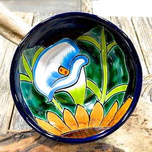 Op de afbeelding: Een blauwe en witte keramische kom met een handgeschilderd ontwerp van een witte en blauwe bloem met groene bladeren en een gele zonnebloem. De kom is perfect om snacks of dips in te serveren.