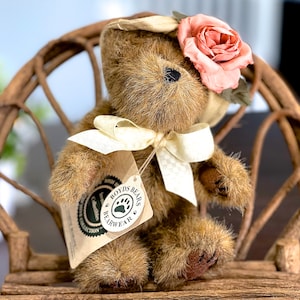 May include: A brown teddy bear wearing a white hat with a pink flower on top. The bear is sitting on a wooden chair and has a white ribbon around its neck. The bear has a tag that says "Boyds Bears Bearwear Collection".