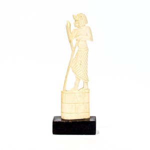 May include: A white ivory sculpture of a person holding a staff, standing on a small wooden base.