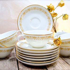 May include: A set of white porcelain teacups and saucers featuring a delicate gold filigree pattern along the rims. The collection includes multiple stacked saucers and several teacups with curved handles, all accented with thin gold trim.