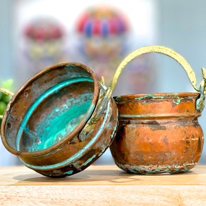 May include: Two small copper pots with brass handles. The pot on the left has a green patina and the pot on the right has a brown patina.