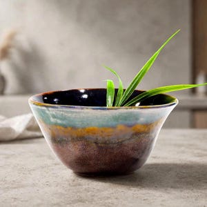 May include: A ceramic bowl featuring a multi-colored reactive glaze with a dark brown base, a band of golden yellow, and a light blue rim. The interior of the bowl is dark, and it contains several long, thin, bright green leaves extending outward.