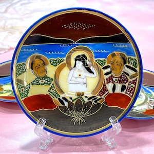 May include: Three blue and gold ceramic plates with a Japanese design. The center plate features a woman in a white robe with a lotus flower. The other two plates feature men in traditional clothing.