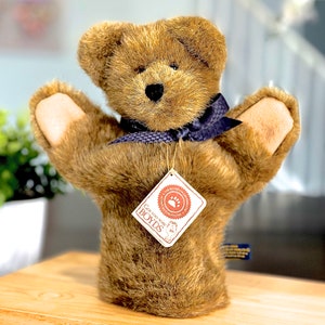 May include: A brown teddy bear hand puppet with a dark blue ribbon around its neck. The puppet has a tag that reads "Boyds". The bear's arms are outstretched, and the fur is a soft, textured material.
