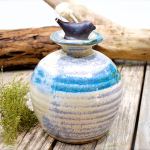 May include: A round ceramic oil lamp with a glossy glaze in shades of blue, white, and lavender. The lamp features a horizontal ribbed texture and a dark, bird-shaped stopper at the top, which holds a thick, white cotton wick for burning.