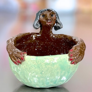 May include: A ceramic bowl with a light green glaze and a woman's face sculpted into the rim. The woman has brown skin, black hair, and red fingernails.