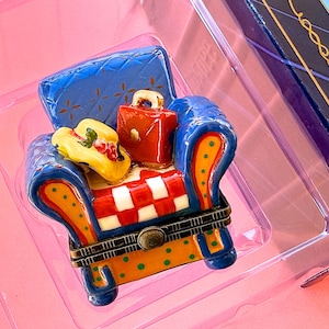 Può includere: Una sedia decorativa in miniatura con schienale e braccioli blu, pois arancioni e seduta a scacchi rossi e bianchi. Un cappello giallo e una valigia rossa sono sulla sedia, suggerendo un tema di viaggio.