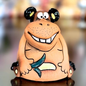 May include: A brown ceramic figurine of a smiling creature with large eyes and a toothy grin. The creature is holding a green leaf in its hands and has a yellow and white design on its belly.