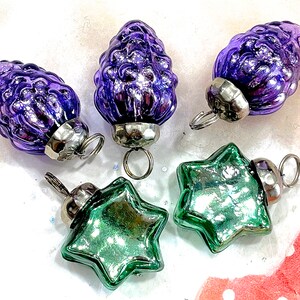 May include: A collection of vintage Christmas ornaments. The ornaments include three purple pinecone-shaped glass ornaments and two green star-shaped ornaments. Each ornament has a silver metal cap and a loop for hanging.